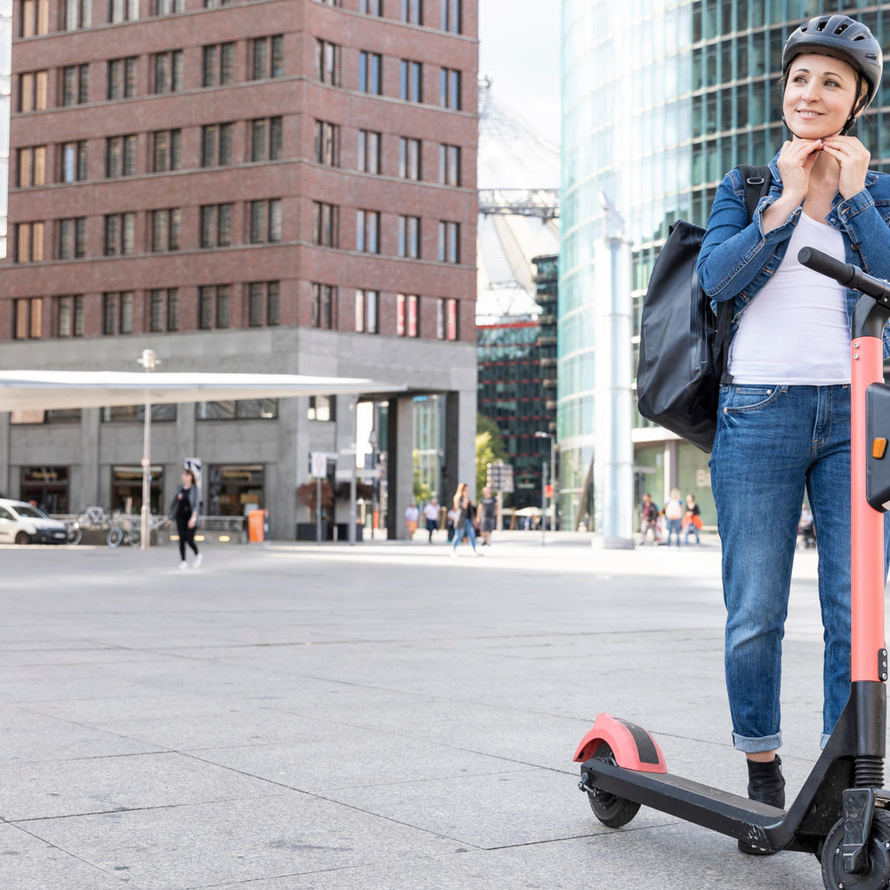 Frau steht neben E-Scooter und setzt sich einen Helm auf.