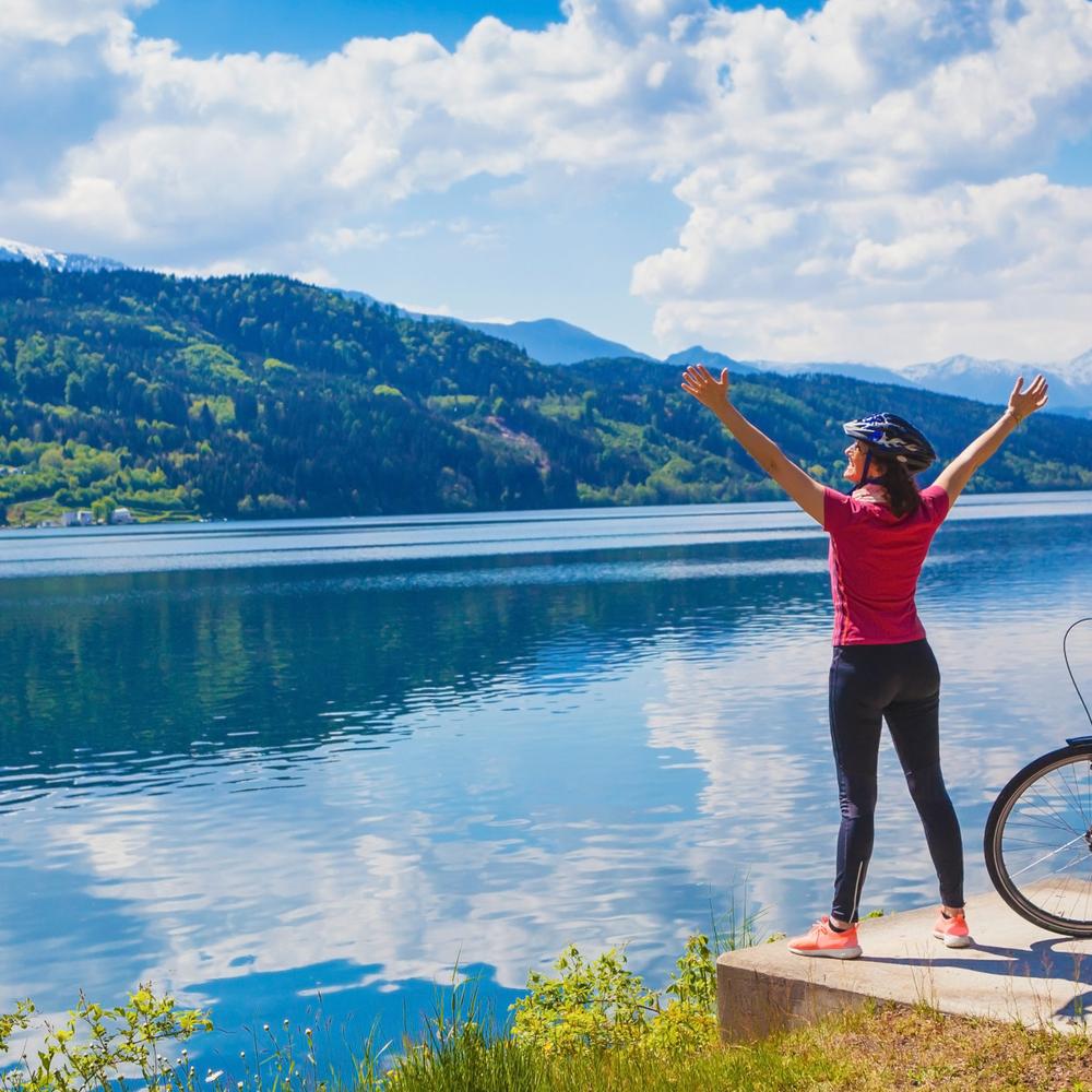 Frau steht vor einem Österreicher See neben einem Fahrrad und hebt ihre Arme freudig nach oben