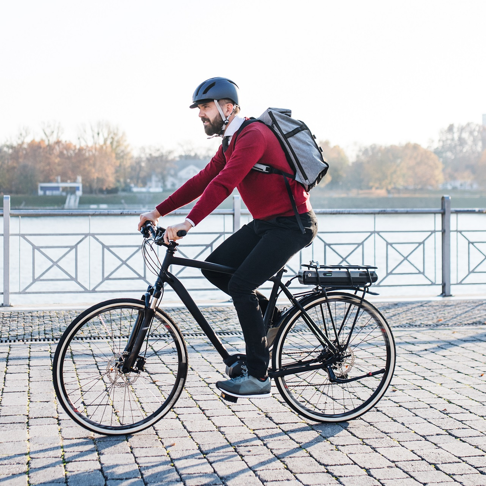 Radfahrer mit Rucksack fährt in städischer Kulisse neben einem Fluss.